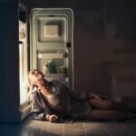 A woman sits on the floor leaning against an open fridge, illustrating real-life use and energy needs for DC vs AC fridges in off-grid living.