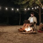 Camper using a laptop by a campfire at night, with string lights and portable gear, illustrating portable power stations for camping.