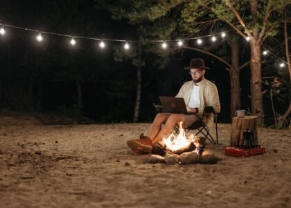Camper using a laptop by a campfire at night, with string lights and portable gear, illustrating portable power stations for camping.