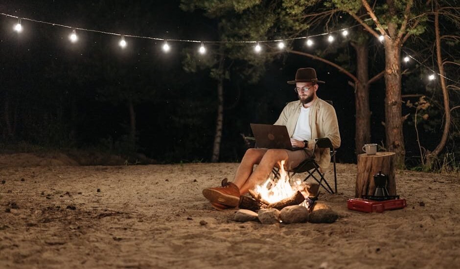 Camper using a laptop by a campfire at night, with string lights and portable gear, illustrating portable power stations for camping.