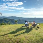 Two campers sit by a table with a portable solar blanket on the grass, overlooking mountains, for a solar blanket review article