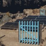 Solar charge controller functions illustrated by ground-mounted solar panels behind a blue gate in a dry, rocky area