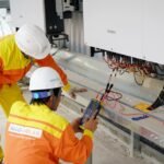 Two technicians in safety gear test a large solar string inverter, illustrating the differences between string and micro inverters.