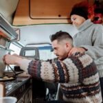 A couple prepares food in a van, showcasing a small 12V freezer in the kitchen area.
