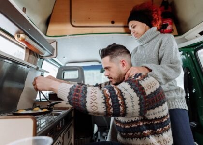 A couple prepares food in a van, showcasing a small 12V freezer in the kitchen area.