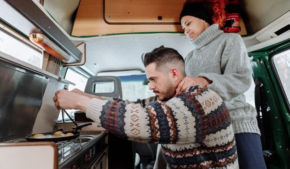 A couple prepares food in a van, showcasing a small 12V freezer in the kitchen area.