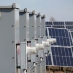 A row of solar combiner boxes next to solar panels, illustrating solar power setup.