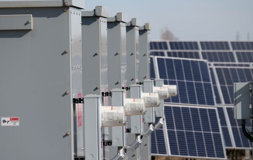 A row of solar combiner boxes next to solar panels, illustrating solar power setup.